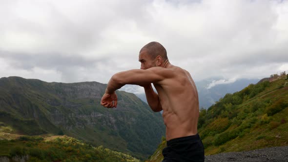 an Unshaven Man with a Bare Muscular Torso is Fighting a Shadow Against the Background of Mountains