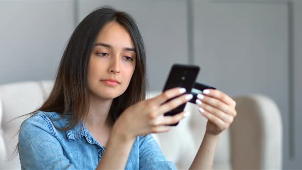 Young Pretty Woman Buying Online with Credit Card and Smartphone Sitting on a Couch in the Living