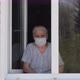 Sad Senior Woman in a Medical Mask Stands at Home at the Window and Look at the Street. - VideoHive Item for Sale