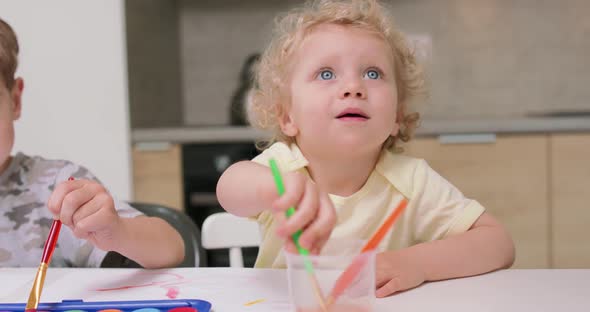 A Little Girl is Mopping a Paintbrush in a Glass of Water Looking Towards the Camera Her Brother's