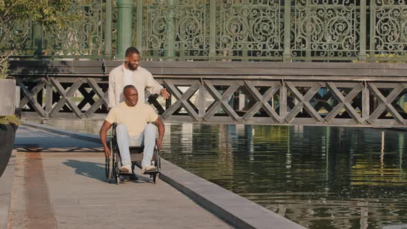 Young Loving Black Guy Accompanies Aged Disabled Person Sitting in Wheelchair During Walk
