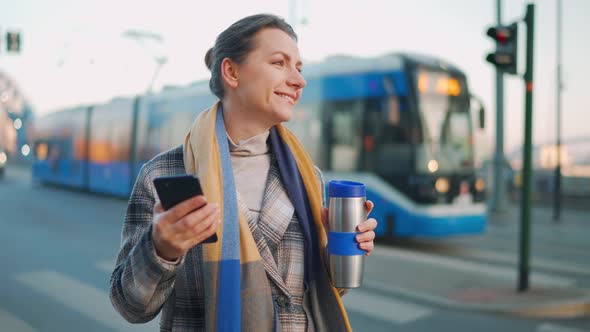 Woman in a Coat Standing on the Street in the Early Morning and Using Smartphone