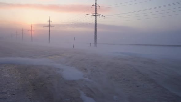 Snow on the Icy Road, the Sun in the Haze. Power Poles Line the Road. Snow Sweeps the Highway in a