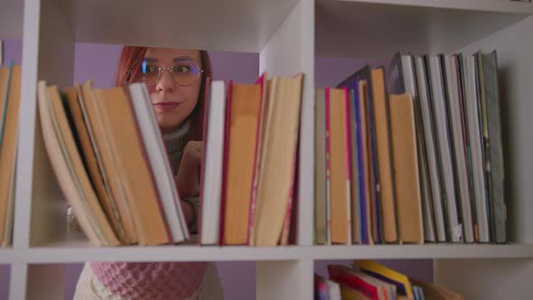 Thoughtful Woman in Glasses Looking at Books Standing Behind Bookshelf