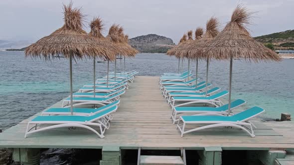 Empty Sun Loungers with Straw Umbrellas in Row on Pier By Beach in Turquoise Sea