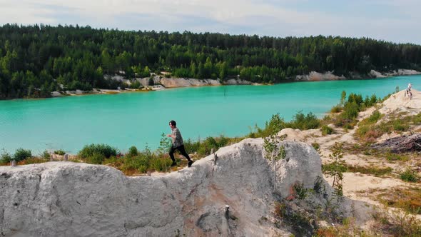 Landscape of a Light Blue Lake Surrounded By Forest - a Successful Man Running on a Rock and Raises