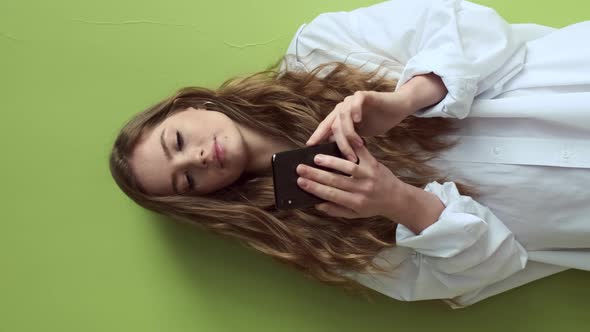 Girl With Long Hair Stands Against Light Green Wall With Mobile Phone In Her Hands.