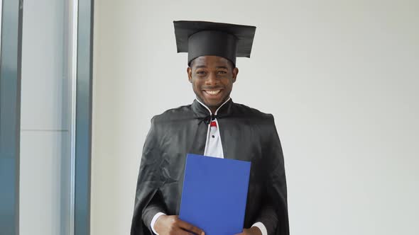 A Young Cheerful AfricanAmerican Graduate Stands in Front of the Camera in a Black Robe and a Master