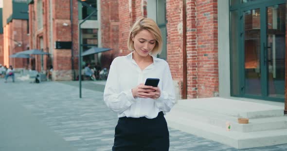 Woman Standing Near Modern Building Typing Text Messages on Smartphone at Nice Day.