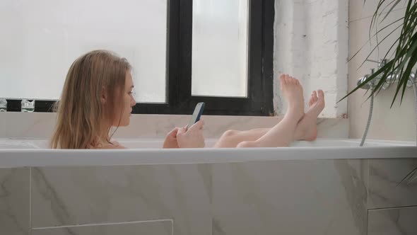 Young Naked Woman Lies in Light White Bathtub and Looks at the Phone. Lady Plays on Cellular in