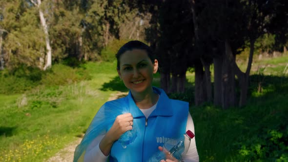 Female Volunteer with a Plastic Bag of Garbage Walks in Front of the Camera and Smiles