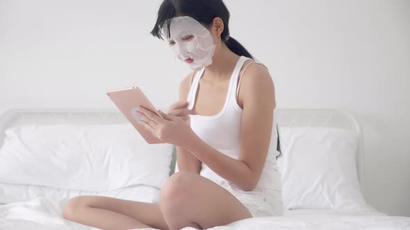 Beautiful young asian woman with sheet facial mask sitting watching tablet computer on bed.