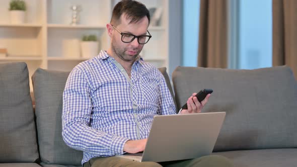 Focused Young Man Using Smartphone and Laptop at Home 