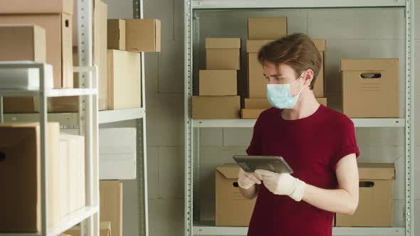 Man Employee of Warehouse in Mask and Gloves Typing on Gadget Tablet Checking or Finding Information