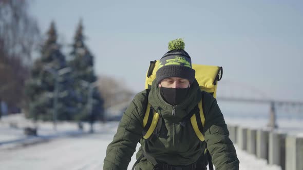 Cyclist with Backpack and Protective Mask Rides Bicycle on Snow Covered Street with Food Delivery