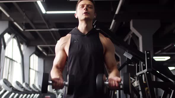 Young muscular man trains in gym lifting dumbbells with two hands, front view. Man of athletic build