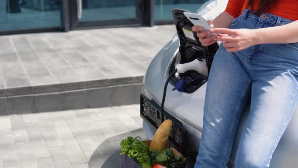 Close Up View of a Girls Uses Phone While Charging Electric Car Near the Shopping Center