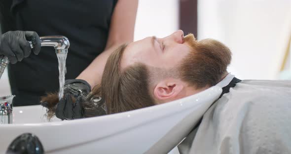 Head of Young Bearded Man Sitting Next to Wash Basin for Washing the Head with Cape and Hands of