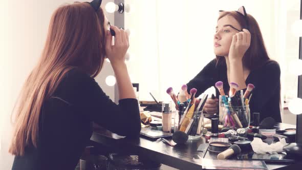 Young Woman Applying Makeup While Sitting at Her Vanity Table with Lots of Cosmetics