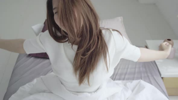 young Asian woman waking up in morning, sitting on bed, stretching in cozy bedroom.