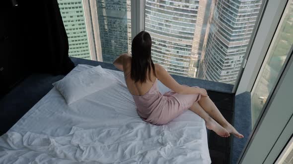 Woman in a Nightgown Lies on a Bed Near a Window in a Corner in a Skyscraper