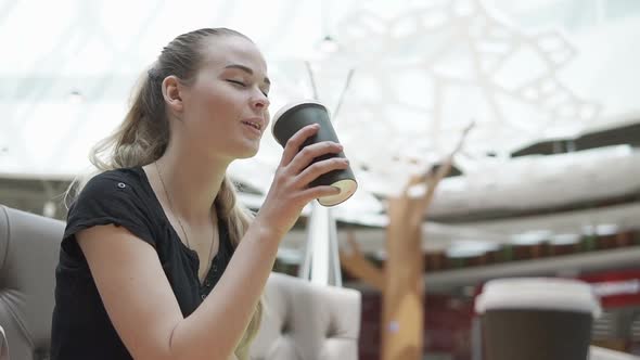 Teenage Girl is Sipping Coffee From Big Cardboard Cup in a Small Cafe Tilt Up