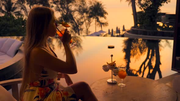 Young Woman Drinks Cocktail Sitting in Luxury Outdoors Restaurant at Hotel or Resort at Evening