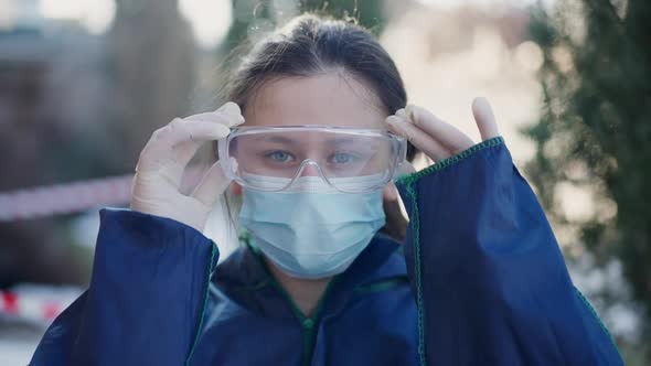 Front View Portrait of Serious Female Criminalist in Face Mask Putting on Protective Eyeglasses