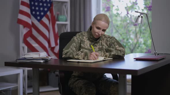 Portrait of Gorgeous Young Woman in Camouflage Uniform Writing Letter Sitting at Table with USA Flag