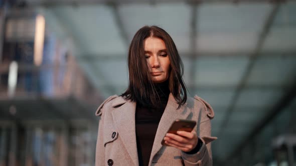 a Brunette in a Gray Coat and with a Phone in Hand Stands Against the Background of a Blurred Glass