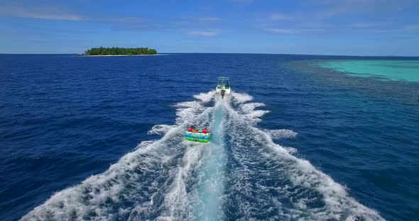 Aerial drone view of man and woman on an inflatable tube towing behind a boat to a tropical island