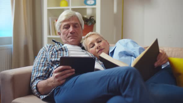 Senior Couple Reading Book and Using Tablet on Couch in Living Room