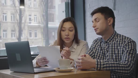 Portrait of a Young Guy and Girl Sitting at a Table with a Laptop Near the Window in a Cozy Cafe or