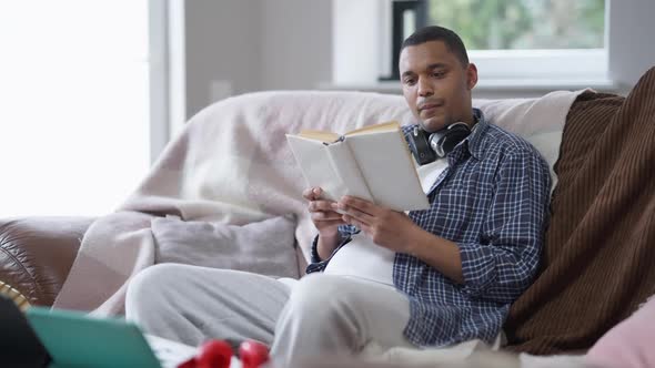 Absorbed African American Man Reading Book Sitting on Couch with Laptop and Tablet on Table