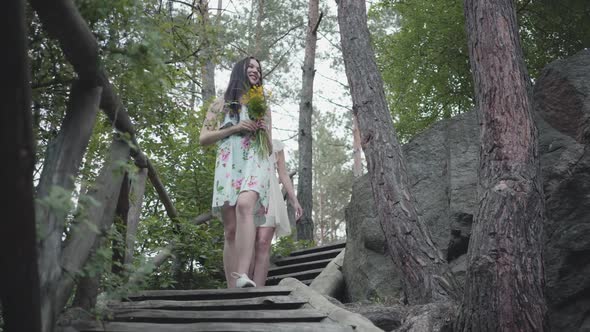 Two Young Women in Short Dreses with Flowers Go Down the Stairs and Looking at Amazing Forest