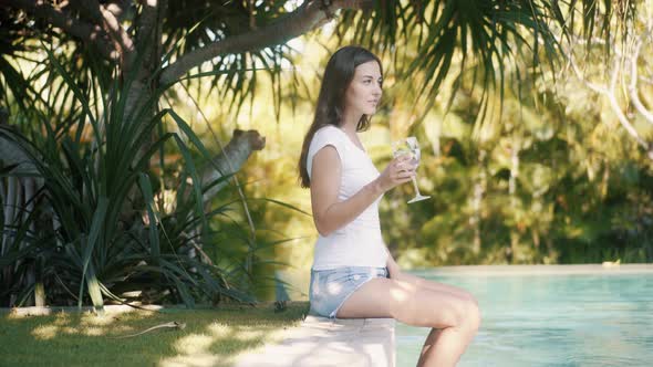 Woman Sits on Edge of Pool Splashes Water with Feet and Drinks Water From Glass