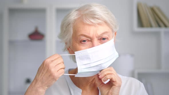 Serious old woman with short grey hair puts on white disposable facial mask