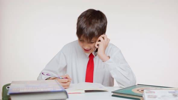 Young Caucasian School Kid in Red Tie Sitting at Table Writing Talking on Mobile Phone on White