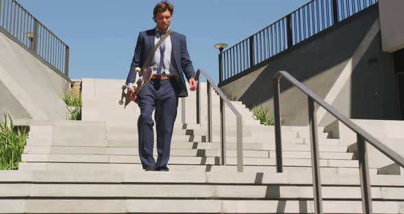 Caucasian businessman walking downstairs, holding skateboard on sunny day