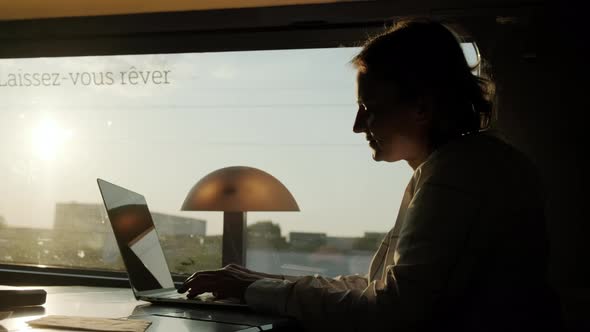 Silhouette of Woman Works on Laptop While Riding Train Sitting Near Window on Sunny Day