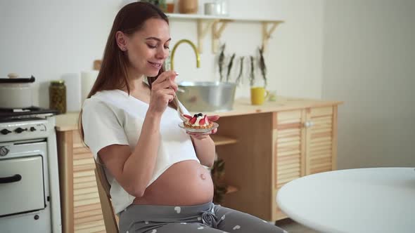 Pregnant Woman Eating Dessert and Smiling While Sitting at Table in Home Room Spbd