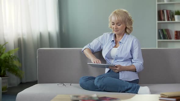 Good-Looking Blonde Senior Woman Sitting on Sofa and Scrolling on Tablet, Apps