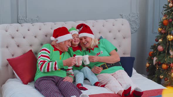 Senior Couple Grandparents Drinking Hot Tea on Bed with Granddaughter Toddler Kid Christmas Morning