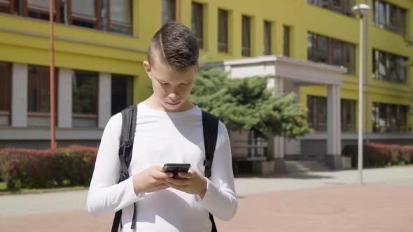 A Caucasian Teenage Boy Works on a Smartphone  a School in the Background