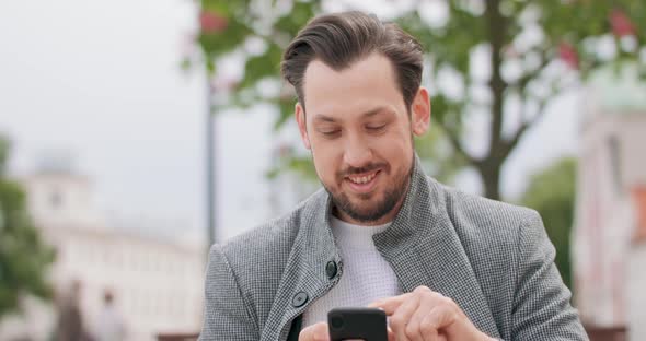 Young Man with Mustaches and a Beard Outside in the Square Smiles Scrolling the Smartphone