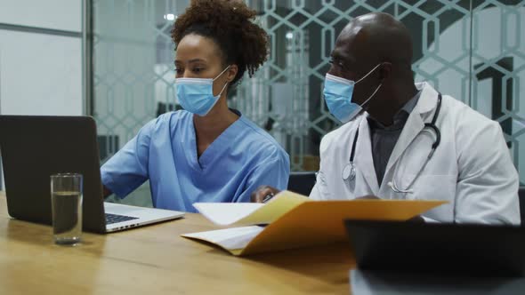 Diverse female and male doctors wearing face masks using laptop and talking in office