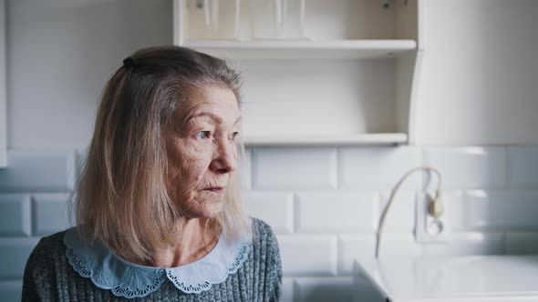 Portrait of Elderly Gray Haired Lady Looking Through the Kitchen Window and Thinking