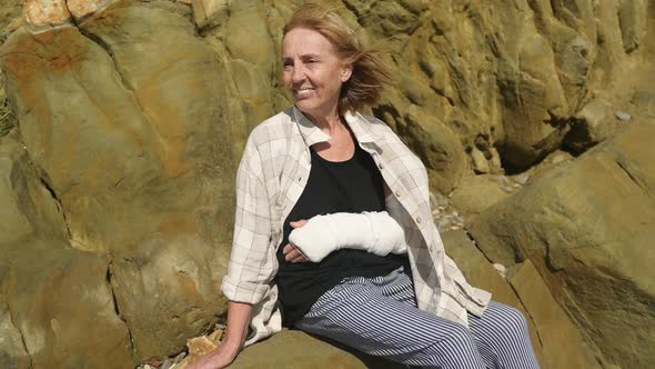 Senior Smiling Elderly Woman Relaxing Sitting on Rocky Coastline in Windy Day Suffering From Joint