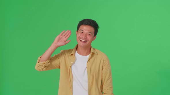 The Happy Young Asian Man Waving Hand While Standing On Green Screen In The Studio