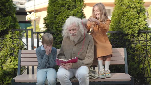 Pretty Caucasian Girl Making Ponytails on Long Grey Hair of Grandfather While Senior Man Reading